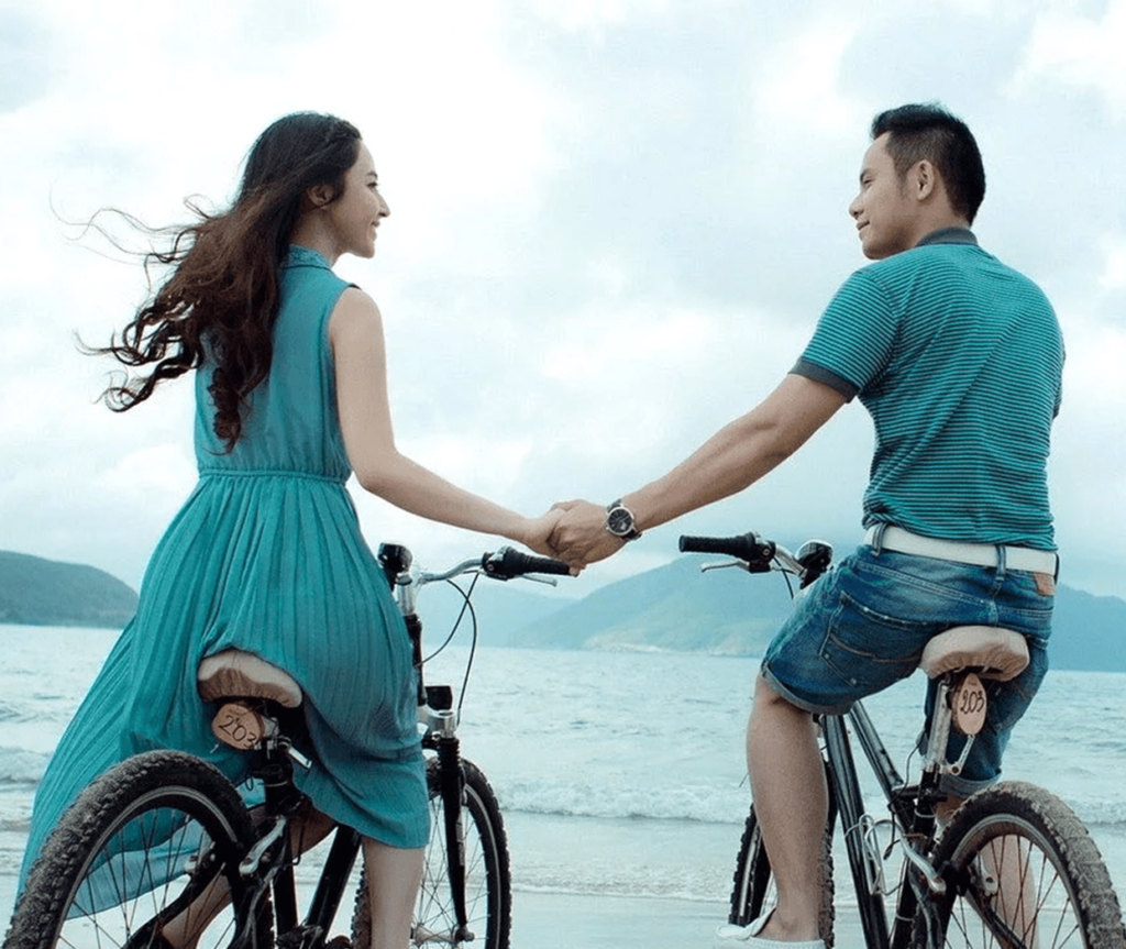 Couples Riding A Bike Whilst Holding Hands Near the Sea